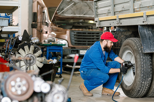 Young technical engineer of car repair service sitting on squats by wheel of large vehicle and drilling